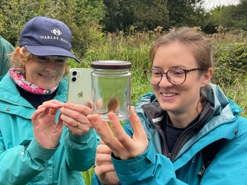 2 women in outdoor clothing examining a harvest mouse in a jar.