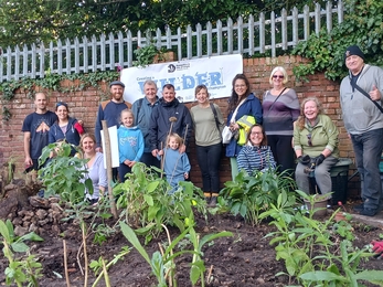 A group shot of 14 volunteers of varying ages. They are standing outside by a flower plant they planted