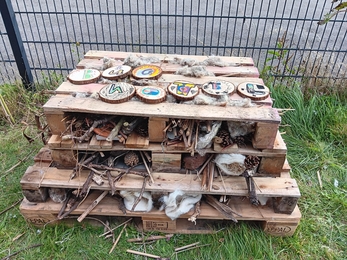 A bug hotel made from pallets, logs and sticks on grass and placed near a metal fence