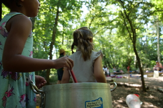 Two children with backs to camera, one child is stirring a spoon in a metal pot. the children are under trees and looking into the distance at other children playing. 