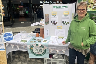 Woman (Amanda) wearing a bright green hoodie next to a table and banner reading 'Wilder Hook'