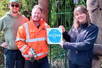 Three individuals standing beside a gate marked "PW" holding a sign that reads "Wilder." The person on the left is dressed in a bright orange work outfit and holding tools, the middle person is wearing a blue jacket and smiling, and the person on the right is holding the sign prominently.