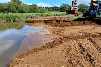 An excavator operates beside a water body, maneuvering over muddy terrain with visible tread marks in a natural setting.