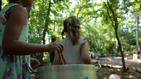 Two children with backs to camera, one child is stirring a spoon in a metal pot. the children are under trees and looking into the distance at other children playing. 