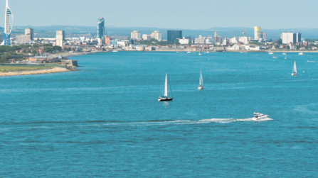 A panoramic view of a coastal cityscape under clear skies. The scene features a body of water in the foreground with two sailboats and a speedboat in motion. The dense urban skyline in the background includes various buildings, with a prominent Spinnaker Tower to the left and tall structures towards the center.