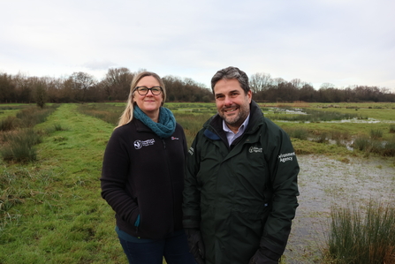 Two individuals wearing black jackets standing outdoors in a grassy, wet field and cloudy sky in the background.