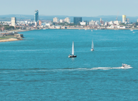 A panoramic view of a coastal cityscape under clear skies. The scene features a body of water in the foreground with two sailboats and a speedboat in motion. The dense urban skyline in the background includes various buildings, with a prominent Spinnaker Tower to the left and tall structures towards the center.